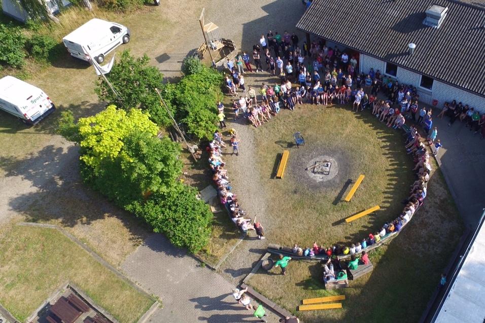 Vogelperspektive auf eine Gruppe von Menschen, die in einem Kreis um ein Lagerfeuer und B&auml;nke sitzen.