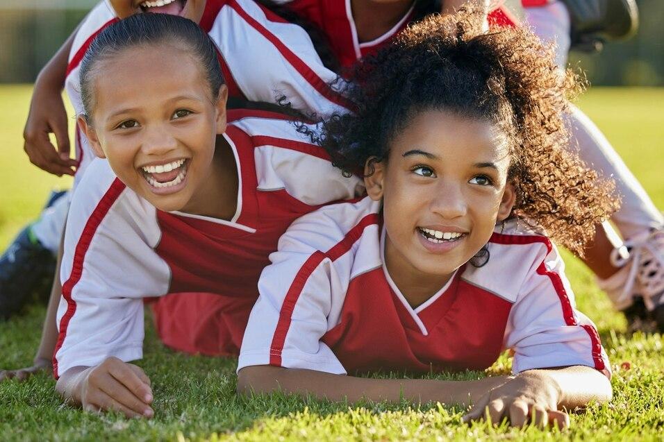 F&uuml;nf junge M&auml;dchen in Fu&szlig;balltrikots l&auml;cheln, w&auml;hrend sie auf einer gr&uuml;nen Wiese &uuml;bereinander liegen.