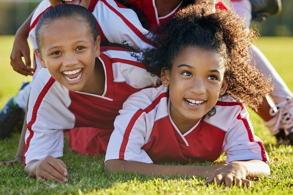 F&uuml;nf junge M&auml;dchen in Fu&szlig;balltrikots l&auml;cheln, w&auml;hrend sie auf einer gr&uuml;nen Wiese &uuml;bereinander liegen.