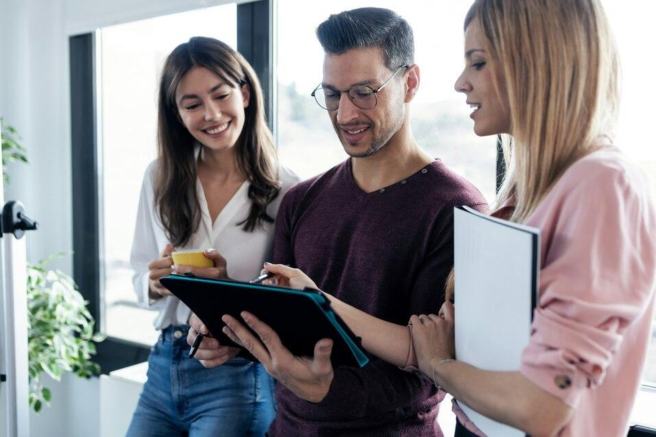 Drei Personen betrachten gemeinsam ein Tablet in einem modernen B&uuml;roraum mit gro&szlig;en Fenstern.