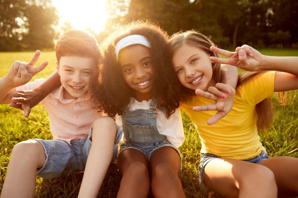 Drei fr&ouml;hliche Kinder sitzen auf der Wiese und zeigen das Peace-Zeichen, umgeben von Sonnenlicht und Gr&uuml;n.