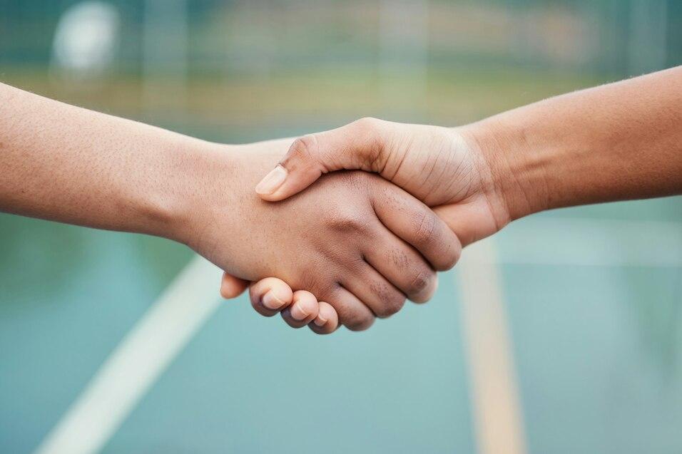H&auml;nde zweier Personen sch&uuml;tteln sich auf einer Sportfl&auml;che, im Hintergrund sind Linien einer Tennisplatzmarkierung sichtbar.