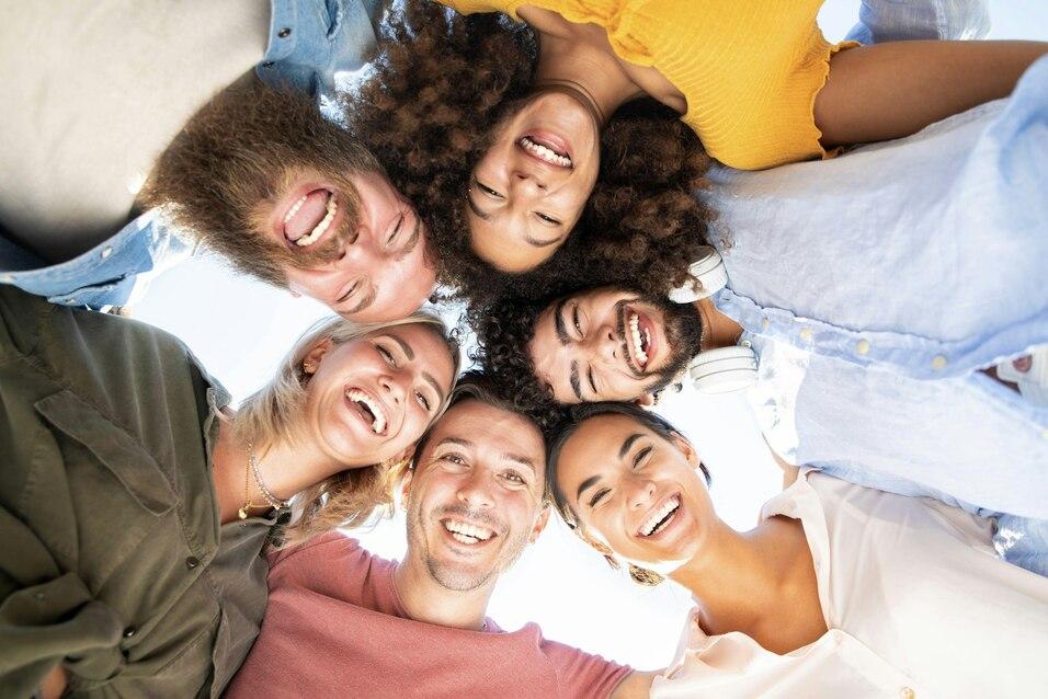 Sechs l&auml;chelnde Menschen umringen die Kamera, die von unten auf ihre fr&ouml;hlichen Gesichter blickt.