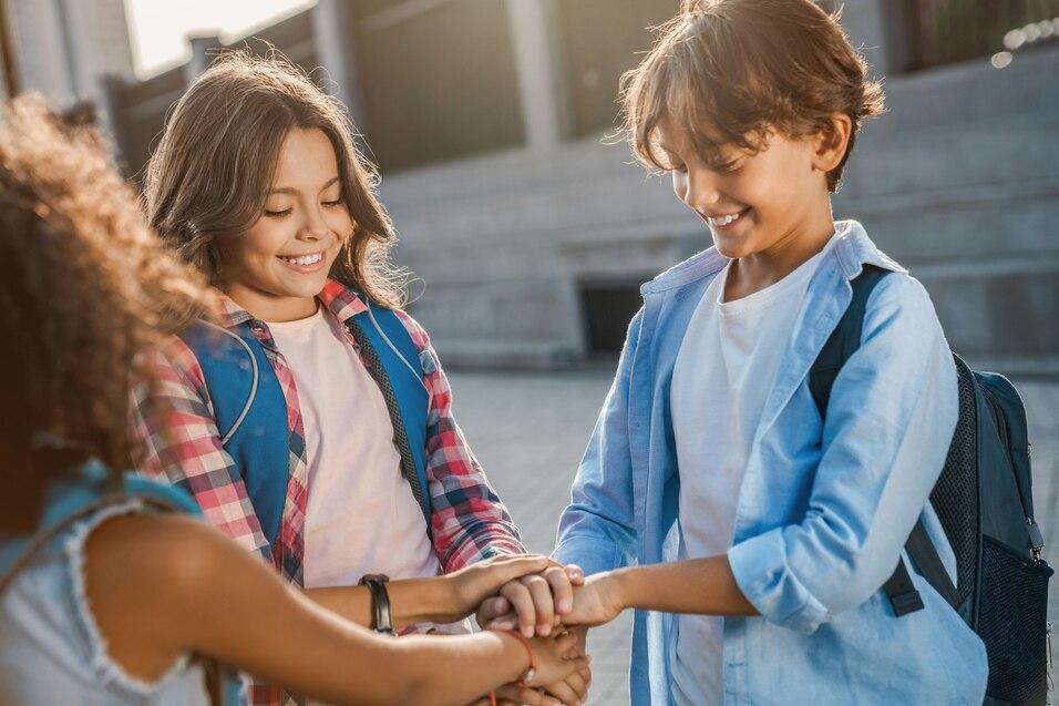 Drei Kinder stehen im Freien und legen ihre H&auml;nde &uuml;bereinander, l&auml;chelnd und in l&auml;ssiger Kleidung mit Rucks&auml;cken.