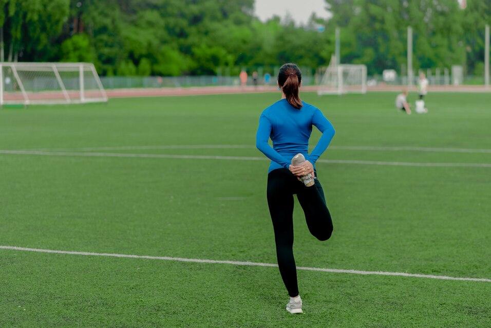Eine Person dehnt im Sportoutfit auf einem gr&uuml;nen Fu&szlig;ballfeld, w&auml;hrend andere im Hintergrund trainieren.