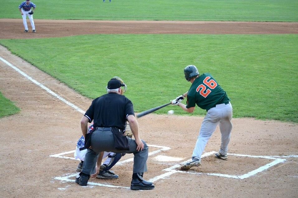 Baseballspieler mit Nummer 26 schl&auml;gt an der Home-Plate, w&auml;hrend der Schiedsrichter hinter ihm steht und ein Spieler im Hintergrund positioniert ist.