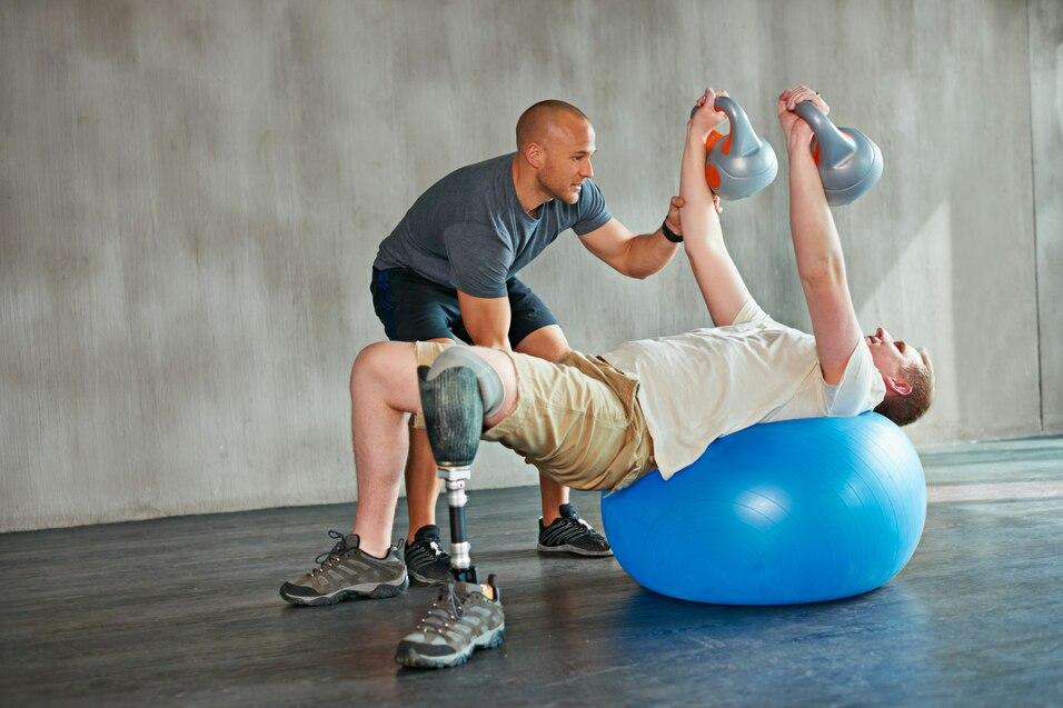 Zwei M&auml;nner trainieren. Einer liegt auf einem Gymnastikball und hebt Kurzhanteln, w&auml;hrend der andere ihn unterst&uuml;tzt.