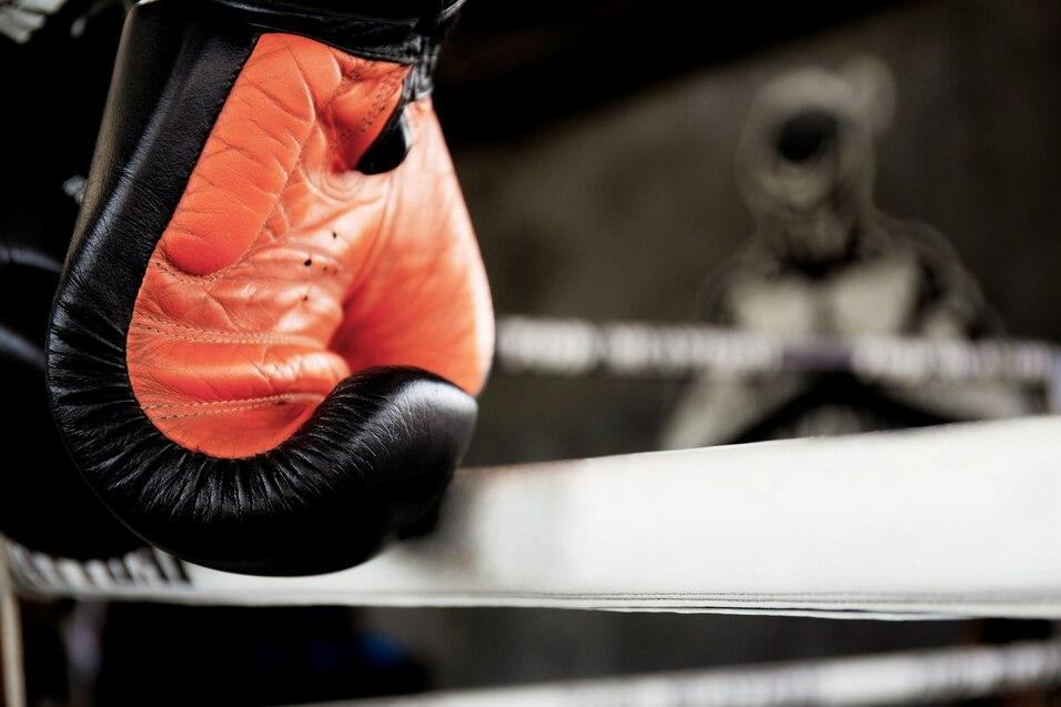 Nahaufnahme eines orange-schwarzen Boxhandschuhs, der an einem Boxringseil h&auml;ngt, mit verschwommenem Hintergrund.