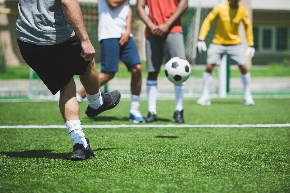 Ein Spieler tritt einen Fu&szlig;ball auf einem Kunstrasenfeld, im Hintergrund stehen weitere Spieler in Sportkleidung.