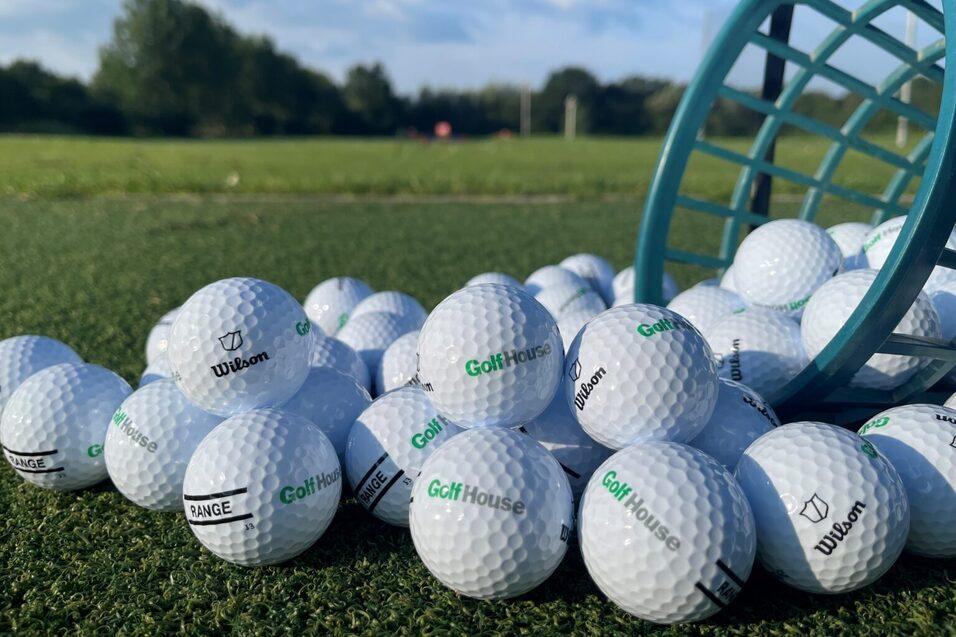 Ein Haufen wei&szlig;er Golfb&auml;lle mit Logo auf einer Golfplatzgr&uuml;nfl&auml;che, einige B&auml;lle liegen neben einem umgedrehten Korb.