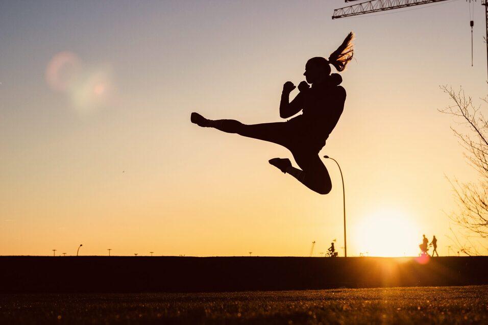 Silhouette einer springenden Sportlerin vor einem Sonnenuntergang mit B&auml;umen und einer Baustelle im Hintergrund.