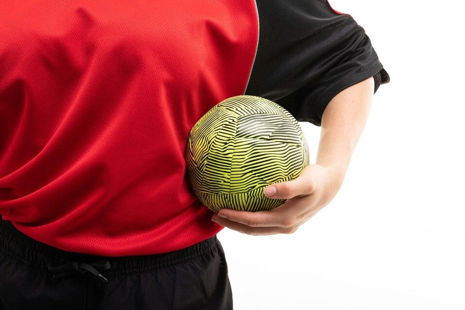 Person in rotem Shirt h&auml;lt einen gestreiften gelben Ball mit einer Hand dicht am K&ouml;rper. Hintergrund ist wei&szlig;.