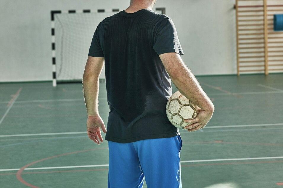 Mann in Sportkleidung steht mit einem Fu&szlig;ball in der Hand auf einem Hallenfu&szlig;ballfeld, Blick Richtung Tor.