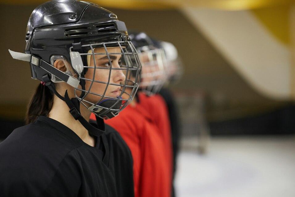 Nahaufnahme einer jungen Eishockeyspielerin im schwarzen Trikot und Helm, die in einer Reihe mit anderen Spielerinnen steht.