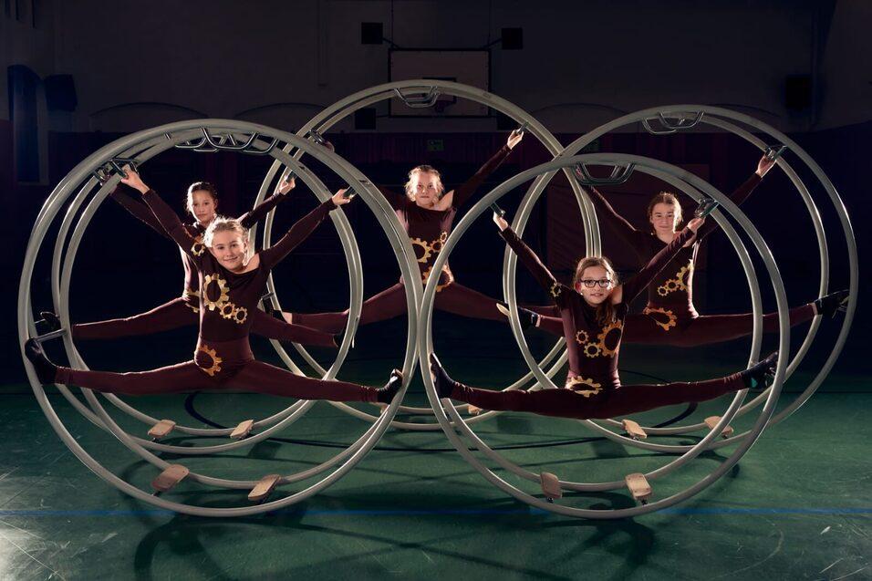 F&uuml;nf Artistinnen in bodenlangen Bodys machen akrobatische Figuren in gro&szlig;en Metallringen auf einem Sportboden.
