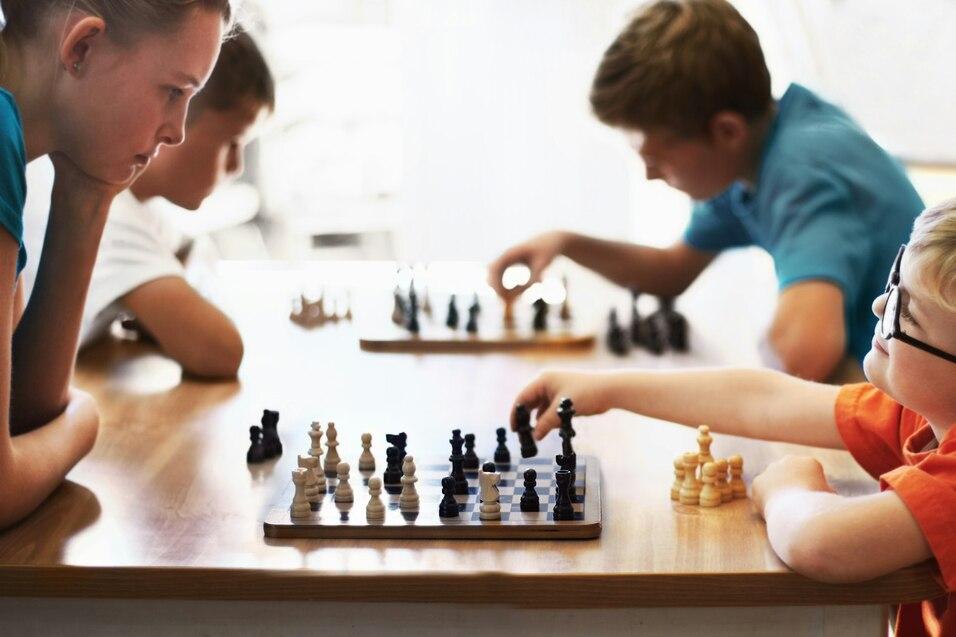 Vier Kinder spielen Schach an einem Tisch, ein Junge bewegt eine Figur, w&auml;hrend die anderen beobachten.