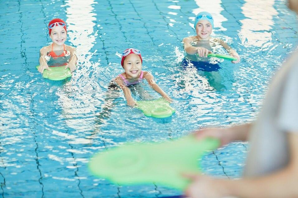 Drei Kinder mit Schwimmbrillen und -kappen schwimmen im Pool, halten gr&uuml;ne Schwimmbretter in den H&auml;nden.