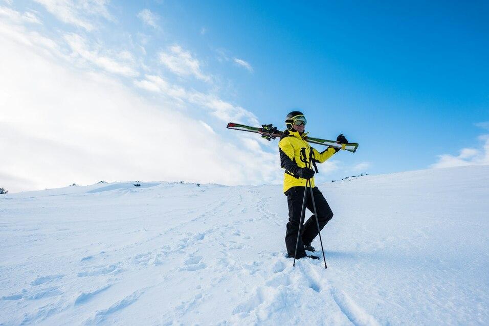 Skifahrer in gelber Jacke steht auf schneebedecktem Gel&auml;nde und tr&auml;gt Skier auf der Schulter, blauer Himmel im Hintergrund.