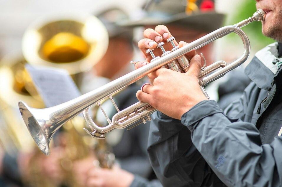 Nahaufnahme einer Hand, die eine silberne Trompete h&auml;lt, mit Musikern im Hintergrund.