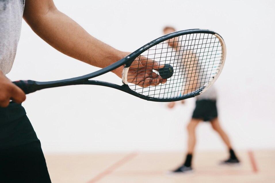 Nahaufnahme einer Hand, die einen Squash-Schl&auml;ger h&auml;lt, w&auml;hrend im Hintergrund ein Spieler auf dem Platz schl&auml;gt.