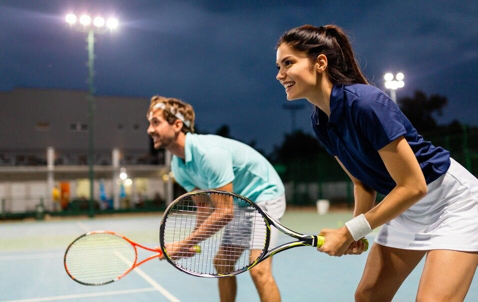 Zwei Tennisspieler, eine Frau und ein Mann, stehen auf einem beleuchteten Platz und bereiten sich auf den n&auml;chsten Schlag vor.