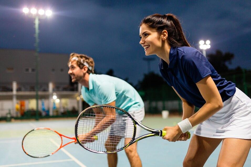 Zwei Tennisspieler, eine Frau und ein Mann, stehen auf einem beleuchteten Platz und bereiten sich auf den n&auml;chsten Schlag vor.