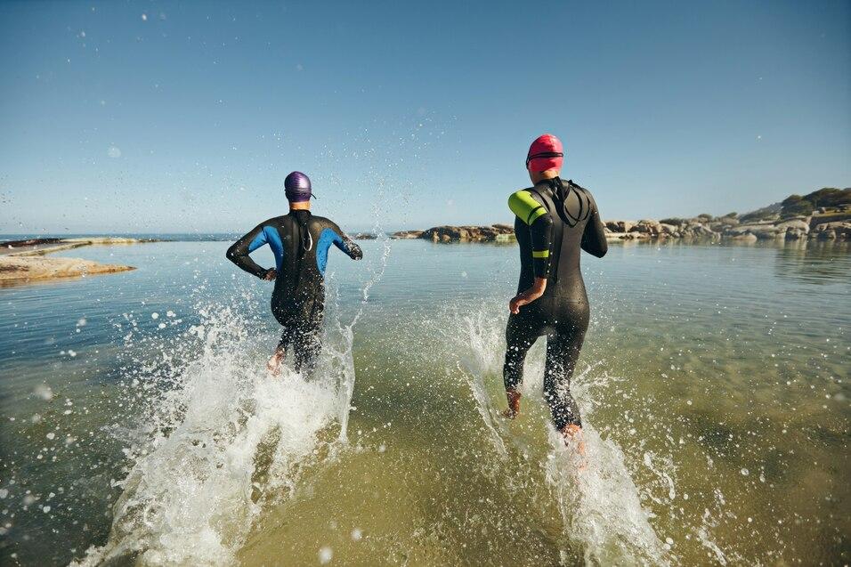 Zwei Schwimmer in Neoprenanz&uuml;gen laufen spritzend ins Wasser bei klarem Himmel.