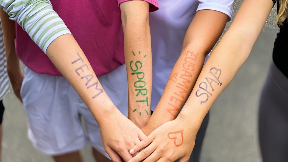 Vier Arme liegen übereinander, mit farbiger Schrift: „TEAM“, „SPORT“, „ZUSAMMEN“, „SPASS“ auf der Haut.