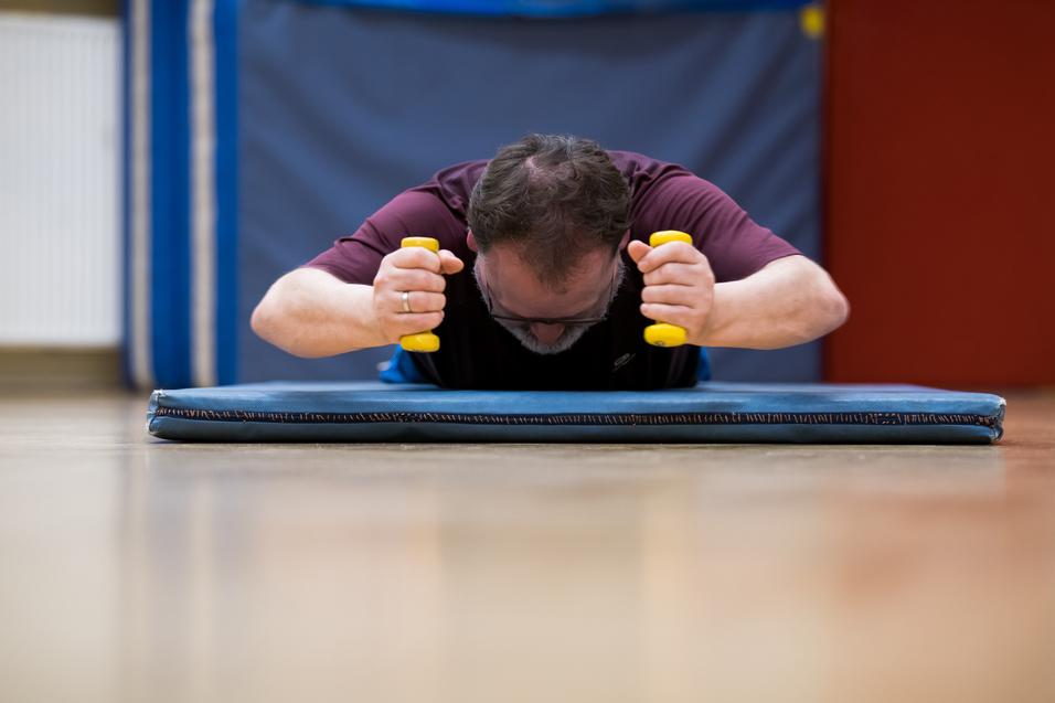 Mann trainiert auf einer Matte mit gelben Hanteln, Kopf nach unten, in einem Sportraum mit blauen Wänden.