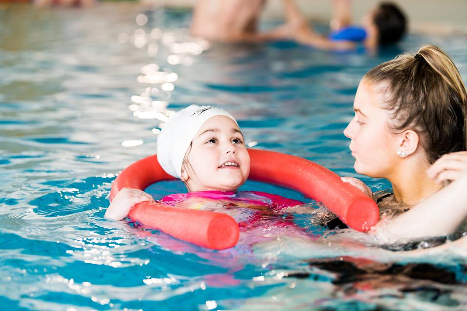 Ein Mädchen mit Schwimmring lächelt in einem Schwimmbecken, während eine Schwimmtrainerin neben ihr steht.